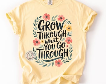 Puede incluir: Camiseta amarilla clara con la frase "GROW THROUGH WHAT YOU GO THROUGH" en negro, rodeada de ilustraciones florales y de hojas rosas, amarillas y verdes. La camiseta tiene mangas cortas y cuello redondo.