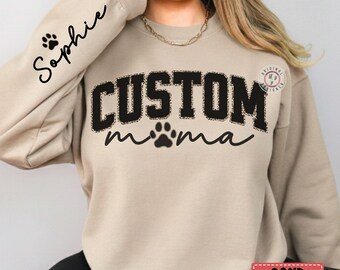 Könnte beinhalten: Sandfarbenes Sweatshirt mit dem Text "CUSTOM mama" in Schwarz, mit einem Pfotenabdruck-Design. Der Ärmel hat den Namen "Sophie" und einen Pfotenabdruck. Das Wort "SAND" befindet sich in einem rechteckigen Feld unten.
