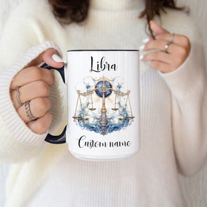 May include: White ceramic mug with a black rim and handle. The mug features a watercolor illustration of a Libra zodiac symbol with a gold scale and blue and white waves. The text "Libra" and "Custom name" are printed on the mug.