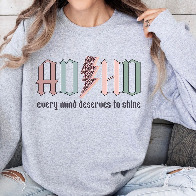 May include: Gray sweatshirt with the words "ADHD" in pink, green, and light blue with a leopard print lightning bolt in the middle. The text "every mind deserves to shine" is below the words ADHD.