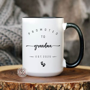 May include: White ceramic mug with a black handle. The mug has black text that reads "Promoted to Grandma Est. 2025" with a pair of footprints below the text.