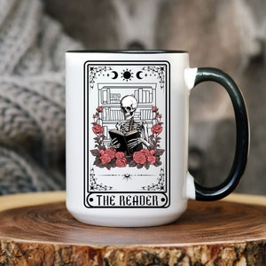 Puede incluir: Taza de cerámica blanca con asa negra. La taza presenta una ilustración en blanco y negro de un esqueleto leyendo un libro frente a una estantería con rosas. El texto "THE READER" está impreso en la taza.