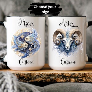 May include: Two white ceramic mugs with black handles. The mug on the left features a blue and gold illustration of two fish with the word "Pisces" above and "Custom" below. The mug on the right features a blue and gold illustration of a ram's head with the word "Aries" above and "Custom" below. The text "Choose your sign" is visible above the mugs.