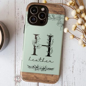 May include: A phone case with a wooden top and bottom, and a light green center. The center features a floral design around the letter "H" and the name "heather" in a script font. The case has a camera cutout.