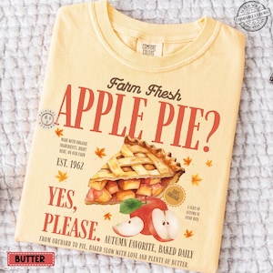 Puede incluir: Camiseta amarillo claro con la frase "Farm Fresh APPLE PIE?" en rojo y marrón. El diseño incluye una ilustración en acuarela de una porción de tarta de manzana, manzanas y hojas de otoño. El texto incluye "Yes, Please" y "Autumn Favorite."
