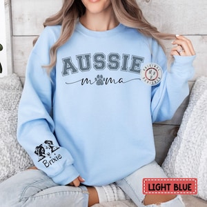 May include: Light blue sweatshirt with the text "Aussie mama" in gray with a paw print. The sweatshirt has a small image of a dog peeking out from the sleeve with the text "Brixie" underneath.