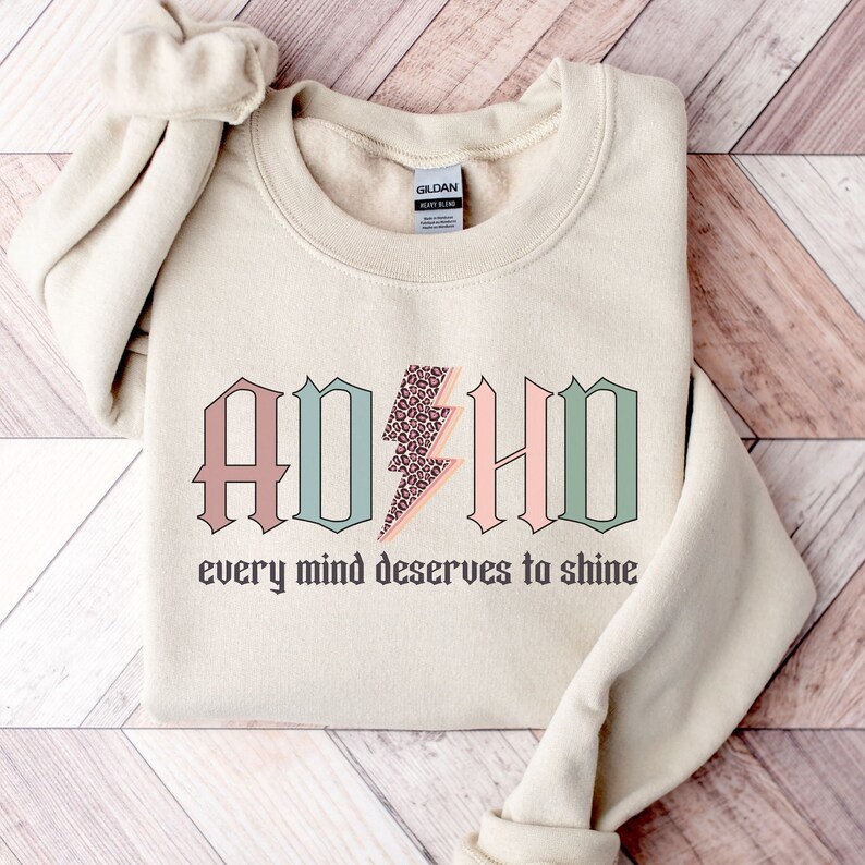 May include: A beige sweatshirt with the words "ADHD" in pink, green, and brown letters with a leopard print lightning bolt in the middle. The text "every mind deserves to shine" is below the letters.