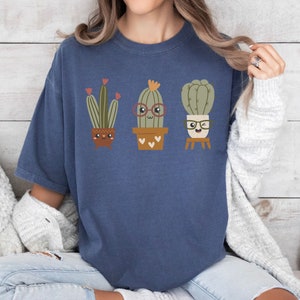 May include: A navy blue t-shirt featuring a graphic of three cartoon cacti wearing glasses. The cacti are in pots and have happy faces.