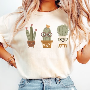 May include: A white t-shirt featuring a graphic of three cartoon cacti in pots. The cacti have faces and wear glasses. The cacti are green, brown, and beige.