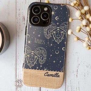 Puede incluir: Funda de teléfono azul marino con un diseño del zodiaco Aries, con una ilustración de carnero, estrellas y lunas. La funda tiene una parte inferior de arpillera con el nombre "Camilla" y el logotipo del diseñador.