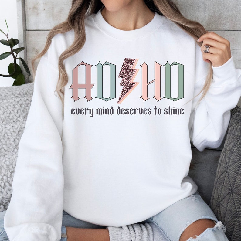 May include: White sweatshirt with a pink, green, and light blue graphic that says "ADHD" with a lightning bolt in the middle. The lightning bolt is filled with a leopard print pattern. The text below the graphic says "every mind deserves to shine".
