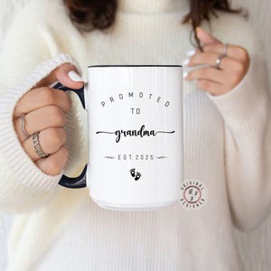 May include: White ceramic mug with a black rim and handle. The mug has black text that reads "Promoted to Grandma Est. 2025" with a pair of black footprints below the text.