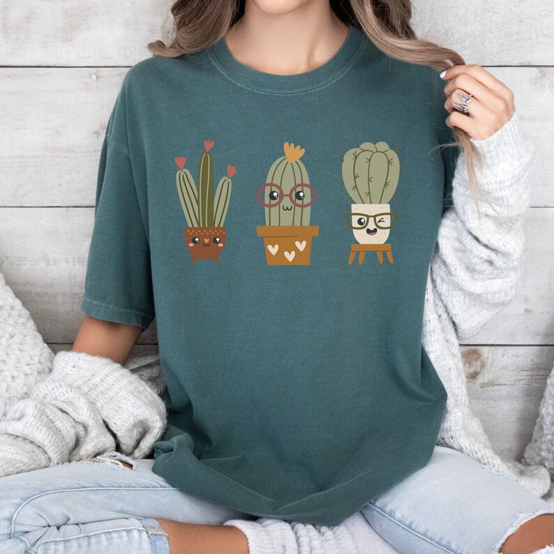 May include: A teal blue t-shirt featuring a graphic of three cartoon cacti wearing glasses. The cacti are in pots and have happy faces.