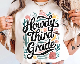 Op de afbeelding: Ivoor kleurig T-shirt met de tekst "Howdy Third Grade" in zwart script. Het ontwerp bevat een cactus, bloemen, een zon, een hoed, een potlood en een rugzak in pastelkleuren. Een casual kledingstuk.