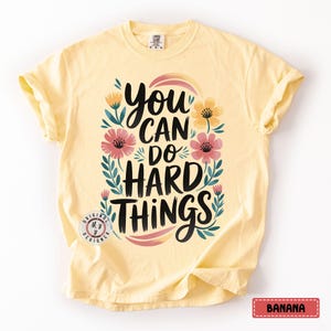 Może przedstawiać: Żółta koszulka z kwiatowym wzorem i napisem "You can do hard things".