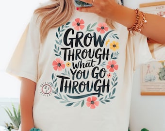 Puede incluir: Camiseta de color marfil con la frase "GROW THROUGH WHAT YOU GO THROUGH" en letras negras. El diseño incluye flores rosas y amarillas y hojas verdes. La camiseta tiene mangas cortas y cuello redondo.