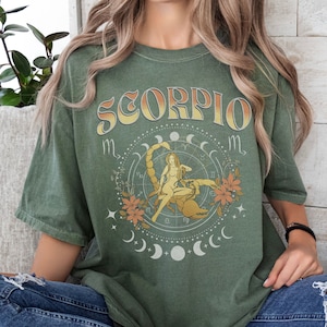 May include: A green t-shirt with a vintage-style graphic of a scorpion and the word "SCORPIO" in a yellow and orange gradient. The graphic also includes a woman sitting on the scorpion, surrounded by stars and crescent moons.