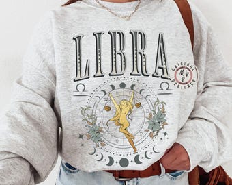 Könnte beinhalten: Ein graues Sweatshirt mit dem Wort "LIBRA" in Schwarzweiß gedruckt. Das Sweatshirt zeigt eine Grafik einer Frau, die eine Waage hält, umgeben von einem Kreis aus Sternen und Monden. Der Text "ORIGINAL M&B DESIGNER" ist in einem Kreis auf der rechten Seite des Sweatshirts gedruckt.