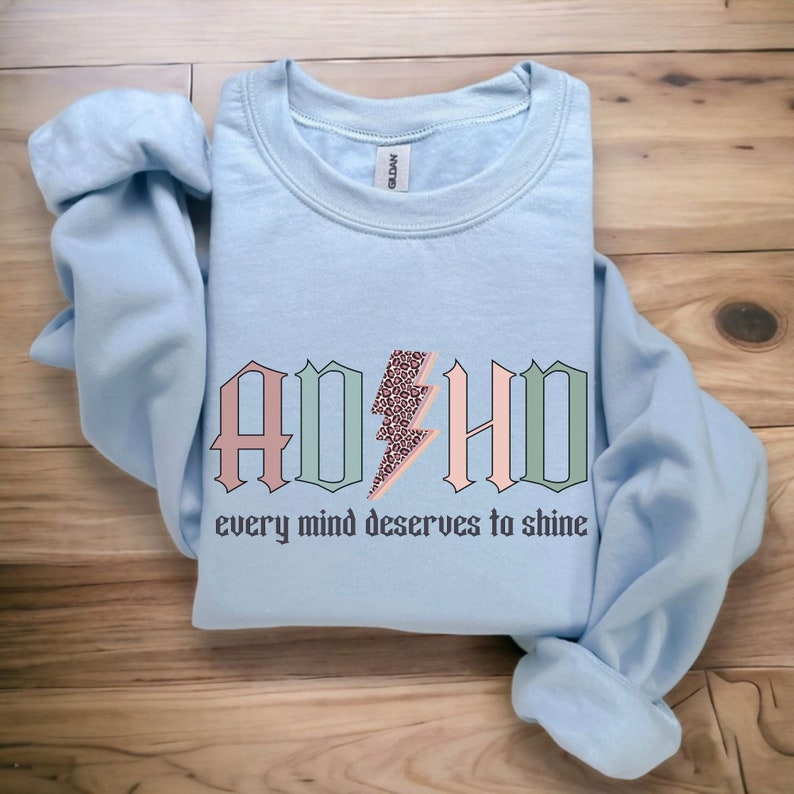 May include: Light blue sweatshirt with the text "ADHD" in pink, green, and white with a lightning bolt graphic. The text "every mind deserves to shine" is below the graphic.