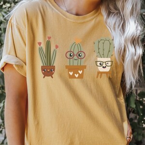 May include: A yellow t-shirt featuring a graphic of three cartoon cacti in pots. The cacti are wearing glasses and have smiling faces.