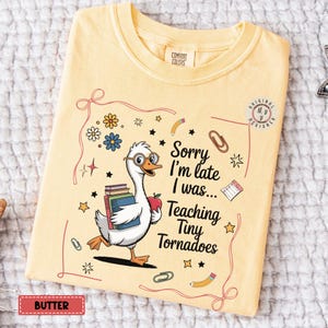 Puede incluir: Una camiseta amarilla con un dibujo animado de un ganso que lleva libros y una manzana. El texto de la camiseta dice "Lo siento, llego tarde, estaba... Enseñando a los pequeños tornados".