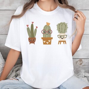 May include: White t-shirt featuring a graphic of three cartoon cacti wearing glasses. The cacti are in pots and have happy faces.
