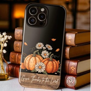 May include: Black phone case featuring a fall design with two orange pumpkins, white daisies, and falling leaves. The case has the text "Give Thanks to the Lord" on a wood-grain background.