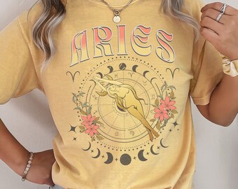 Puede incluir: Una camiseta amarilla con un gráfico de estilo vintage del signo zodiacal Aries. El gráfico presenta a una mujer con el pelo suelto, una rueda celestial y detalles florales. La palabra "ARIES" está impresa en letras grandes y en negrita sobre el gráfico.