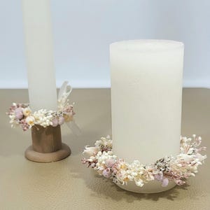 Könnte beinhalten: Zwei weiße Kerzen mit getrockneten Blumenkränzen um die Basis. Die Kerzen stehen auf Holzständern.