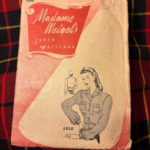 May include: A vintage Madame Weigel's paper pattern envelope, number 1058, featuring a red and beige design. The front displays a black and white illustration of a long-sleeved button-up shirt with chest pockets and a cinched waist, along with text.