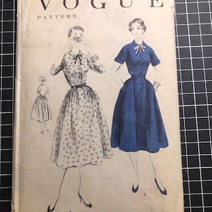 Peut inclure: Enveloppe de patron Vogue vintage avec des illustrations de deux robes, une bleue et une à motifs floraux. L'enveloppe est vieillie, avec le texte "Vogue Pattern" et des informations de taille. Daté de 1954.