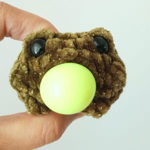 May include: A handmade, brown crocheted frog with black plastic eyes and a light green ball for a mouth. The frog is made of a textured yarn with gold flecks. The frog is a whimsical, handmade item.