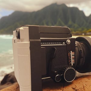 Pode incluir: Uma câmera de médio formato preta e prateada com uma lente e um rótulo "PROFESSIONAL S" na frente. A câmera está sentada em uma superfície marrom com uma praia e montanhas ao fundo.