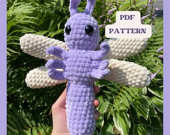 May include: A crocheted dragonfly plush toy with a purple body and white wings. The toy is held in a hand and the text "PDF PATTERN" is visible in the background.