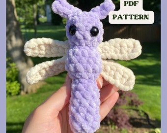 May include: A crocheted dragonfly toy with a purple body and cream colored wings. The toy is made with a fuzzy yarn and has large black eyes. The text "PDF PATTERN" is visible in the image.
