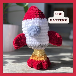 May include: Crochet pattern PDF for a red, white, and gray rocket toy. The rocket has a round nose, two red wings, and a yellow and orange base.
