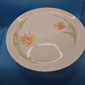 May include: A white, round serving platter with a floral design. The platter features two yellow flowers with orange centers and green leaves. A thin, orange and yellow line curves around the flowers. The platter is on a blue background.