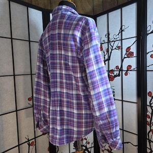May include: A long-sleeved button-down shirt featuring a plaid pattern in shades of purple, blue, and white. The garment has a standard collar, buttoned cuffs, and a curved hemline, displayed on a mannequin to show the back and side profile.
