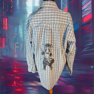 May include: Gray and white plaid button-down shirt with a black graphic on the back. The graphic is a cartoon character wearing a hat and sunglasses, holding a drum and a drumstick. The shirt has long sleeves and a classic collar.