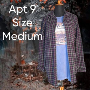 May include: A long-sleeve button-down shirt with a purple and dark gray plaid pattern worn over a blue t-shirt with graphic text. The text on the shirt reads Apt 9 Size Medium and includes a graphic with the text LAG M Since 1998 NHAT.