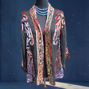 May include: Black open-front jacket with velvet floral patterns in red, purple, and pink. The jacket features gold embroidered trim along the edges and a blue beaded necklace. The jacket is displayed on a black mannequin.