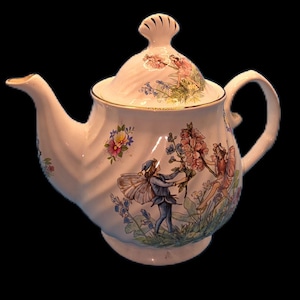May include: A white porcelain teapot with a ribbed, swirling surface and a shell-shaped lid handle. The body features colorful illustrations of winged fairies interacting with large pink flowers and green foliage, accented by gold trim along the rim.