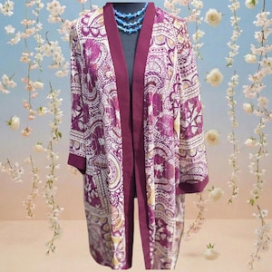 May include: A long, open-front kimono-style robe with a vibrant paisley pattern in shades of purple, white, and gold. The robe features a deep burgundy trim along the edges and sleeves. A blue beaded necklace is visible at the neckline.