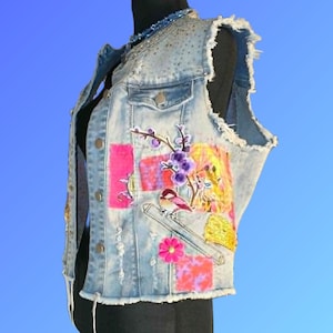 May include: A light blue denim vest featuring a frayed armhole and hem, embellished with colorful fabric patches, a bird illustration, and a floral motif. The garment includes metallic buttons, distressed detailing, and a beaded neckline trim.