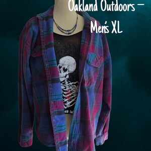 May include: A long-sleeve button-up flannel shirt in a plaid pattern of purple, blue, and dark green. Underneath is a black t-shirt featuring a white skull graphic with heart-shaped eyes and a red heart. Text reads Oakland Outdoors - Men's XL.