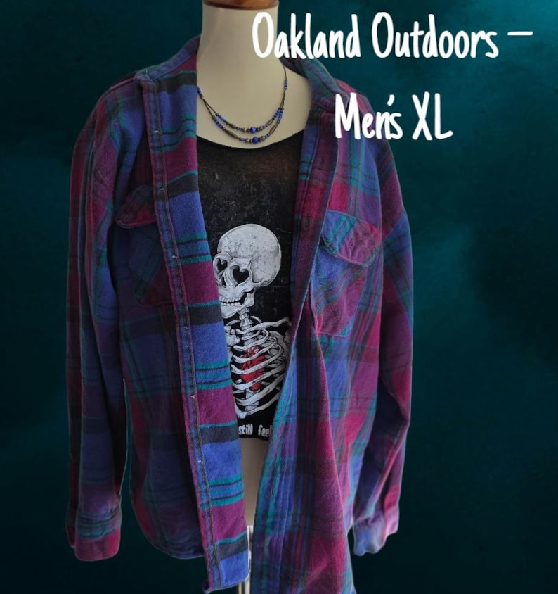 May include: A long-sleeve button-up flannel shirt in a plaid pattern of purple, blue, and dark green. Underneath is a black t-shirt featuring a white skull graphic with heart-shaped eyes and a red heart. Text reads Oakland Outdoors - Men's XL.
