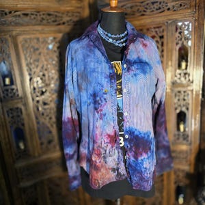 May include: A tie-dye button-down shirt in shades of blue, purple, and red, with vertical white stripes. The shirt is displayed on a mannequin, layered over a black t-shirt and a blue beaded necklace.