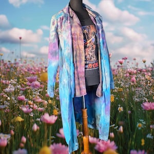 May include: A long-sleeved denim duster coat featuring a tie-dye pattern in shades of blue, purple, and pink. Underneath, a plaid button-down shirt is layered over a black t-shirt with a graphic print featuring Japanese text and figures.