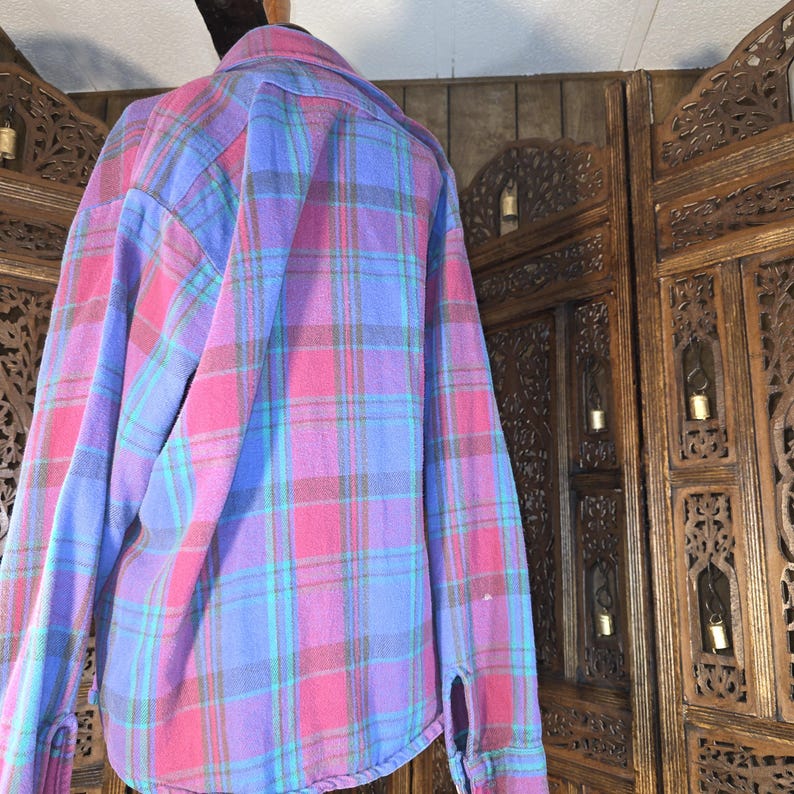 May include: A long-sleeved button-down shirt featuring a plaid pattern in shades of bright pink, light blue, and dark grey. The shirt has a standard collar, a buttoned front placket, and buttoned cuffs on the sleeves, displaying a soft, brushed texture.