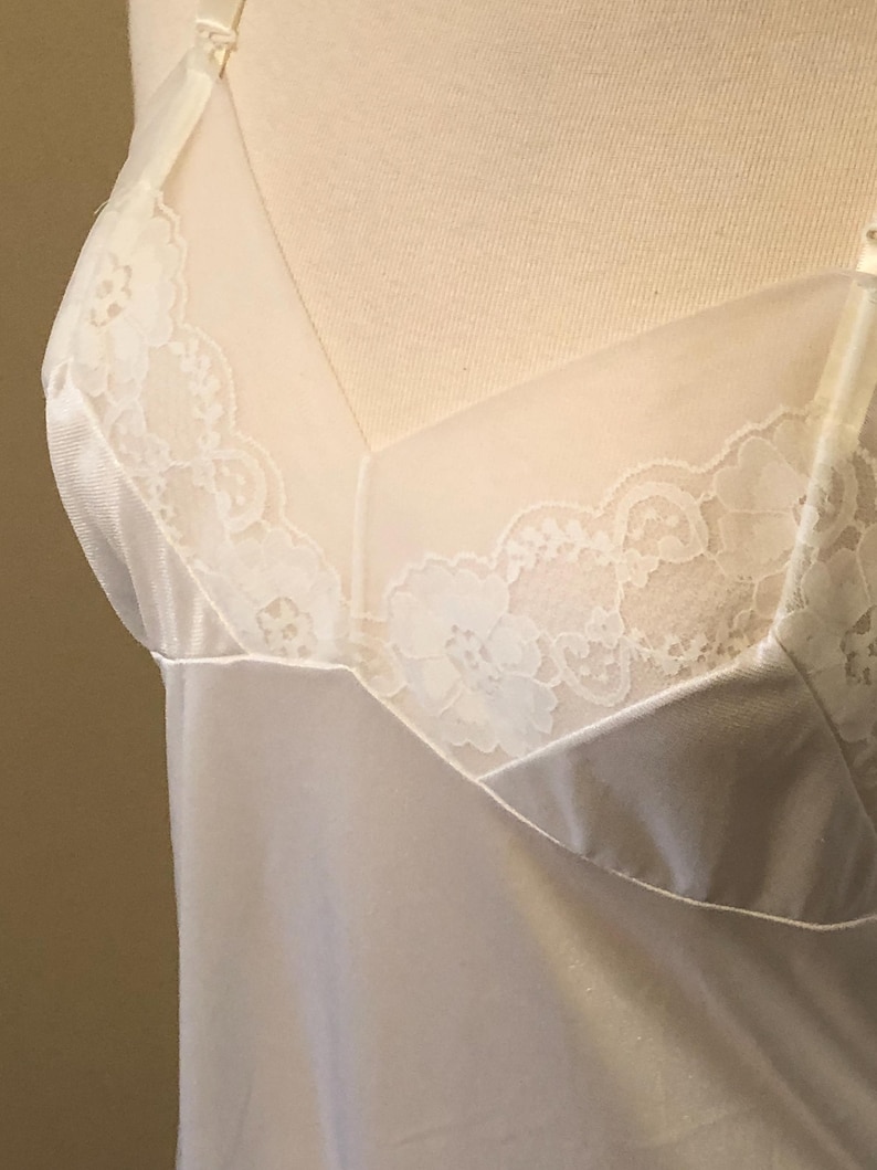 May include: Cream-coloured lingerie with delicate lace detailing. The slip features a satin-like fabric and a sheer panel at the neckline, adorned with floral lace. The straps are thin and adjustable.